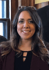 Brittany McDougal Bialy: A woman with long brown hair, wearing a burgundy blazer and black top, smiles at the camera indoors with glasses resting on her head.