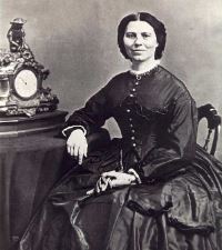 Clara Barton in a dark, long-sleeved dress is seated next to an ornate clock on a table, facing the camera with a neutral expression. The image is in black and white.