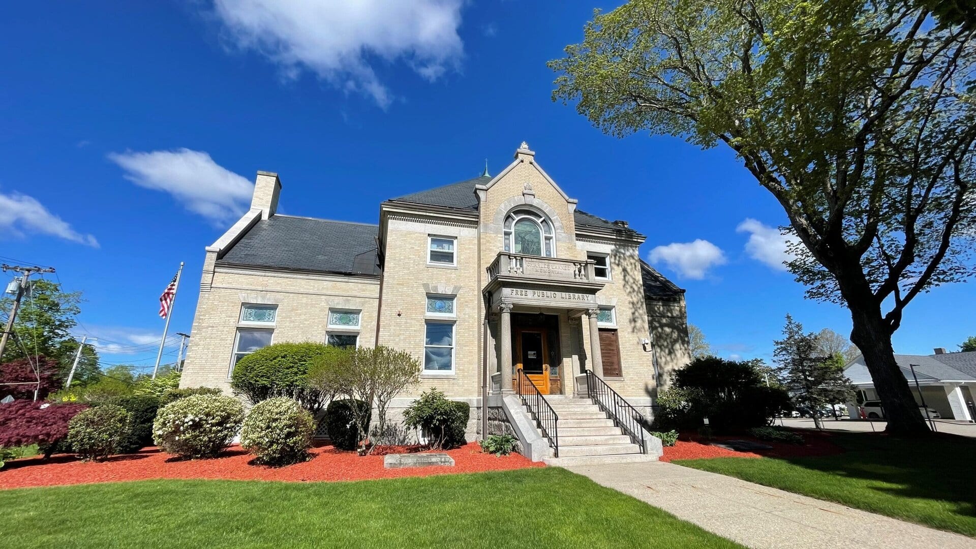 a two story stone building with steps leading to the entrance, surrounded by manicured bushes, grass, and a large tree, under a partly cloudy blue sky.