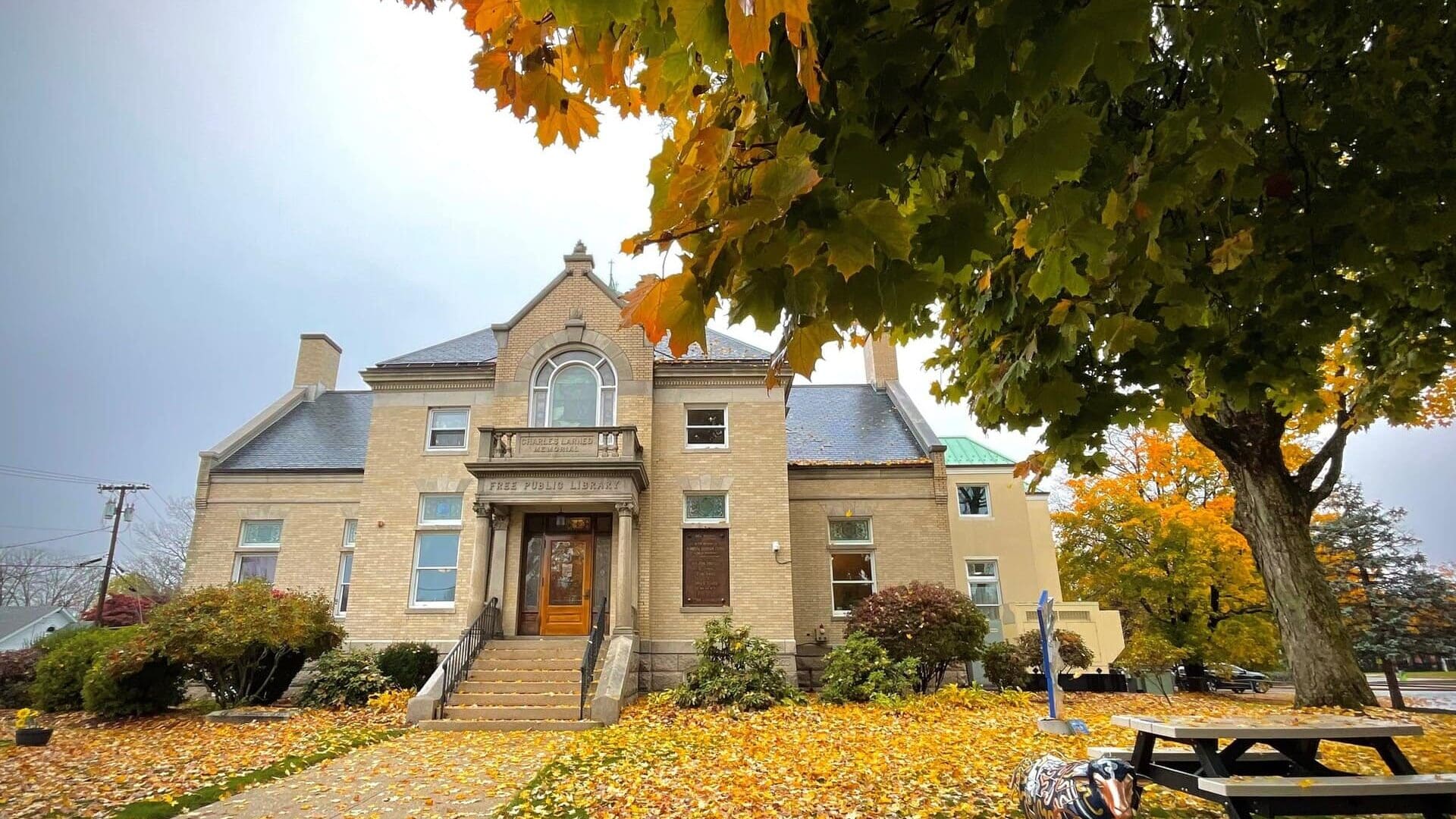 a brick library building with steps leading to the entrance, surrounded by autumn trees and fallen leaves, with a picnic table and a painted cow sculpture in front.