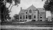 Black and white photo of a large, two-story brick building with a central entrance, wide steps, and surrounding trees.