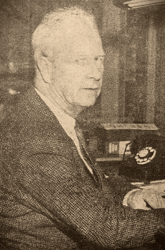 Francis G. Howarth: Black and white photo of an older man in a suit sitting at a desk beside a rotary telephone, looking toward the camera.