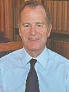 Timothy A. Kelley: A man in a light blue shirt and dark tie is smiling, seated in front of shelves filled with books.
